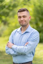 Headshot of a young white man in his 30s standing in front of green trees.