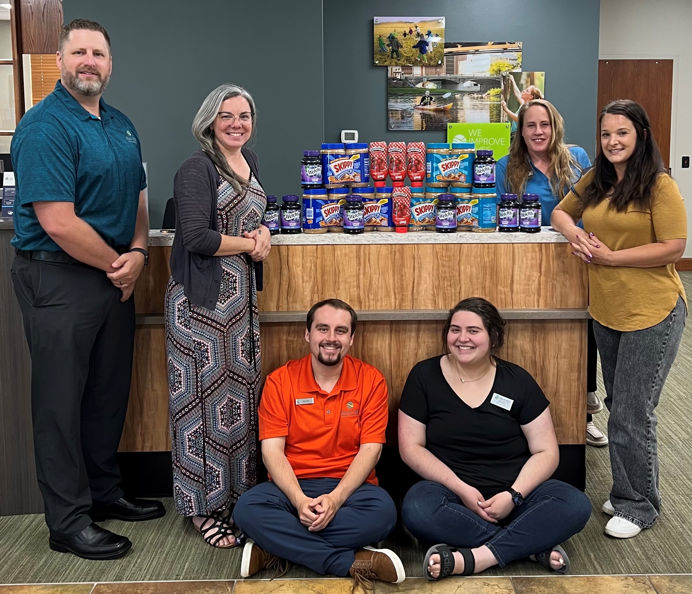 Two men and four women stand and sit around desk displaying peanut butter and jelly donations.